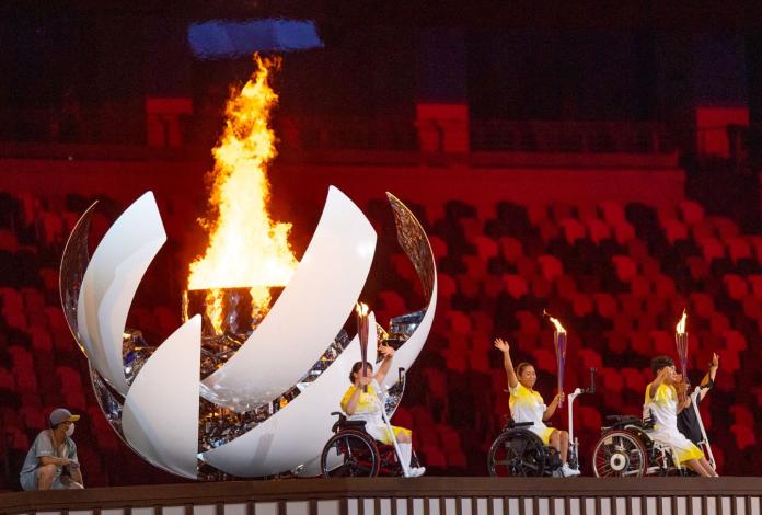 Three athletes in wheelchairs are each carrying the Tokyo 2020 torches in front of a sphere-shaped cauldron